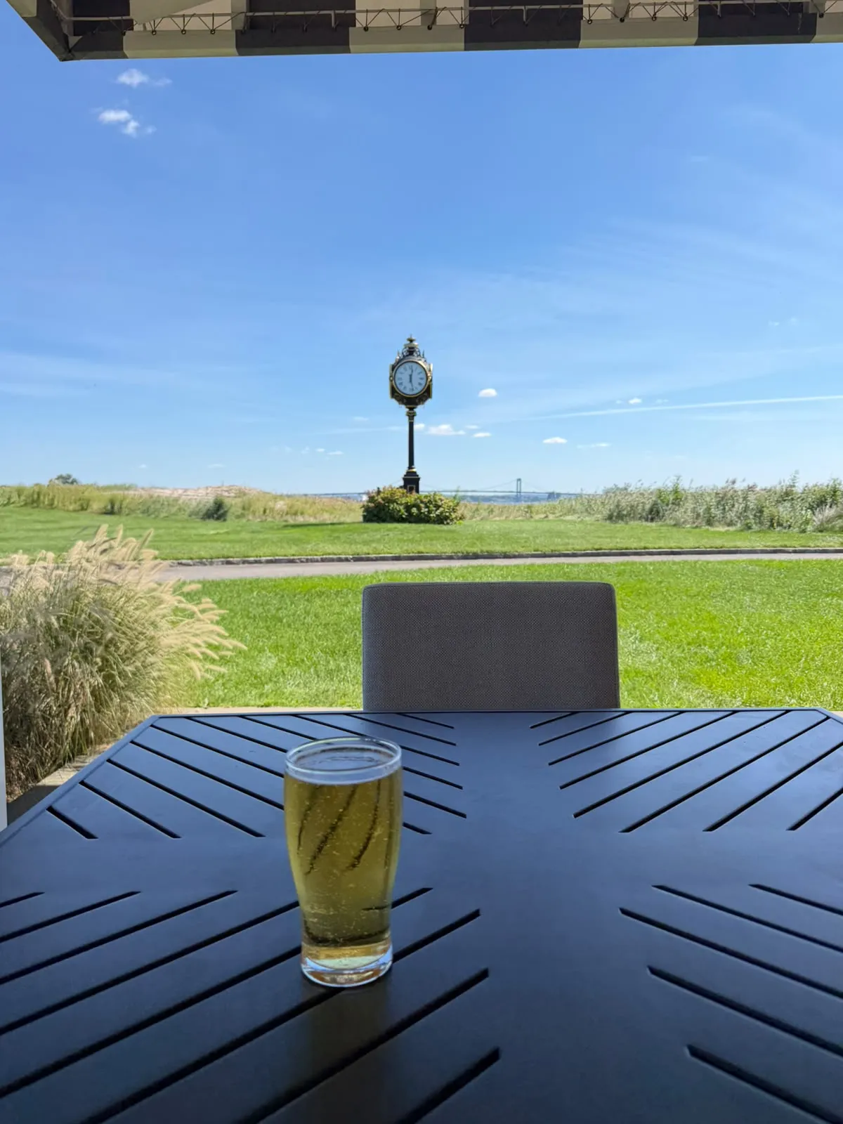 A glass of lager on a dark patio table looking out across a lawn to an ornate street clock, with a suspension bridge on the horizon.