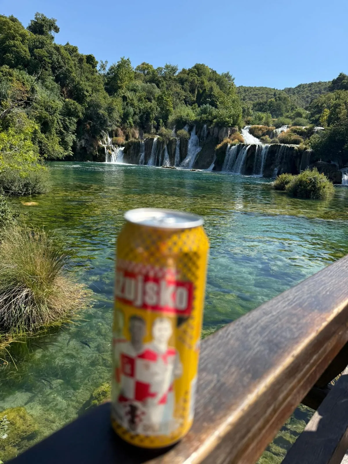 A yellow Ožujsko can standing on a wooden railing above a clear turquoise river, with a wide run of low waterfalls and dense green woodland behind.