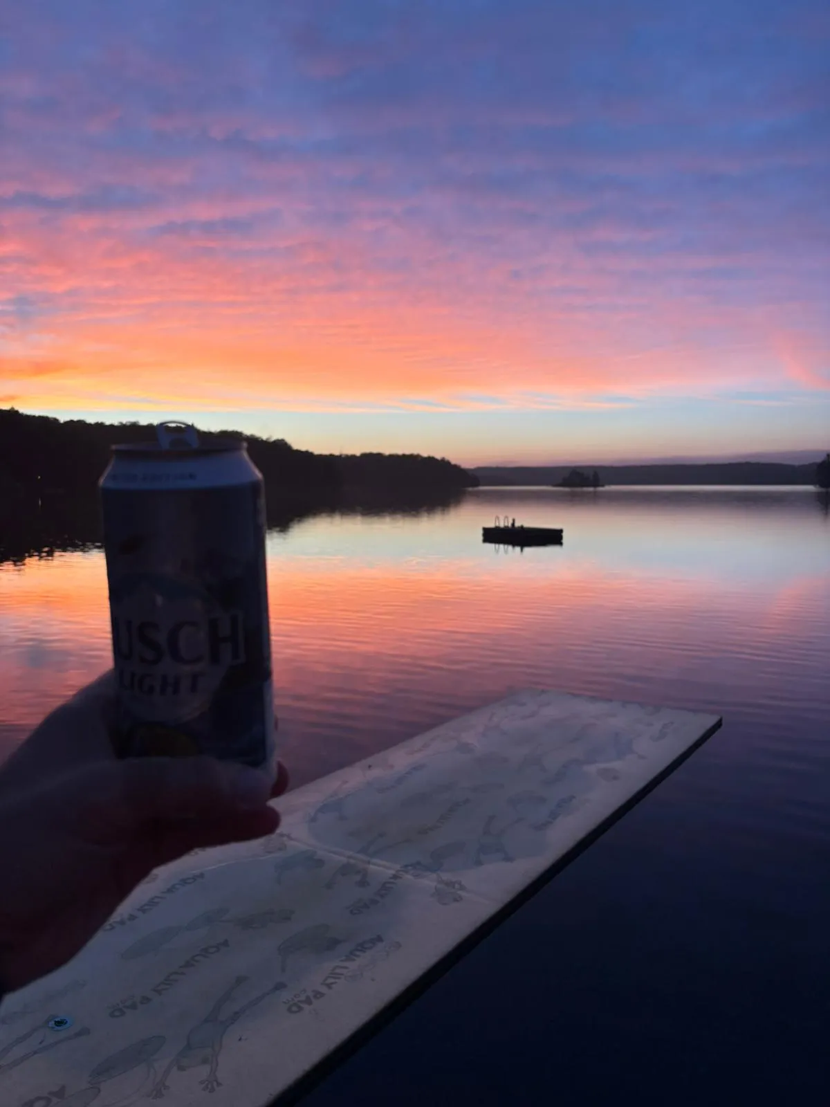 A Busch Light can held up against a still lake at sunset, pink and orange cloud reflected in the water with a swim raft moored offshore.