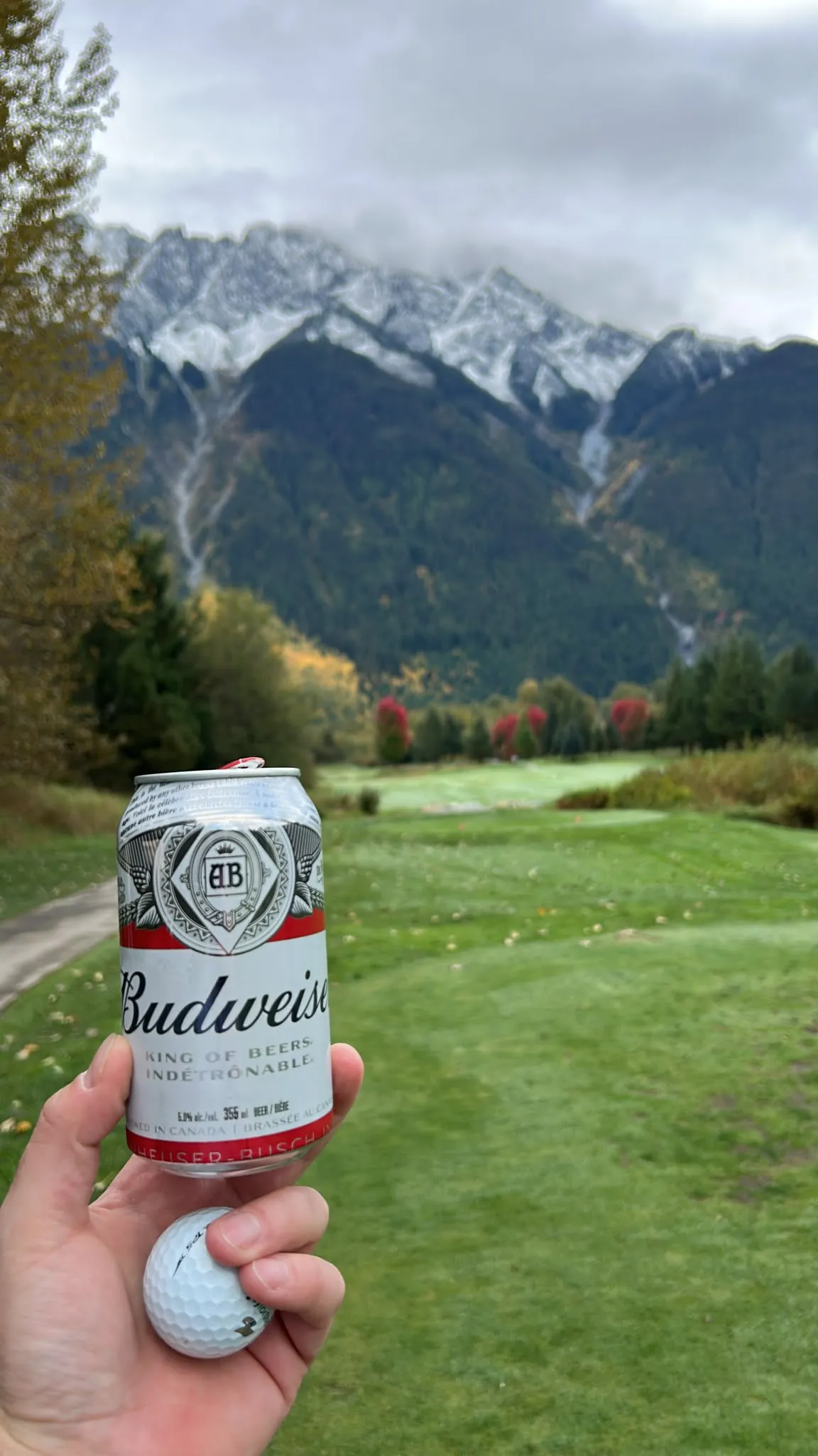 A Budweiser can and a golf ball held up on a fairway, autumn trees and snow-dusted mountain peaks rising behind.