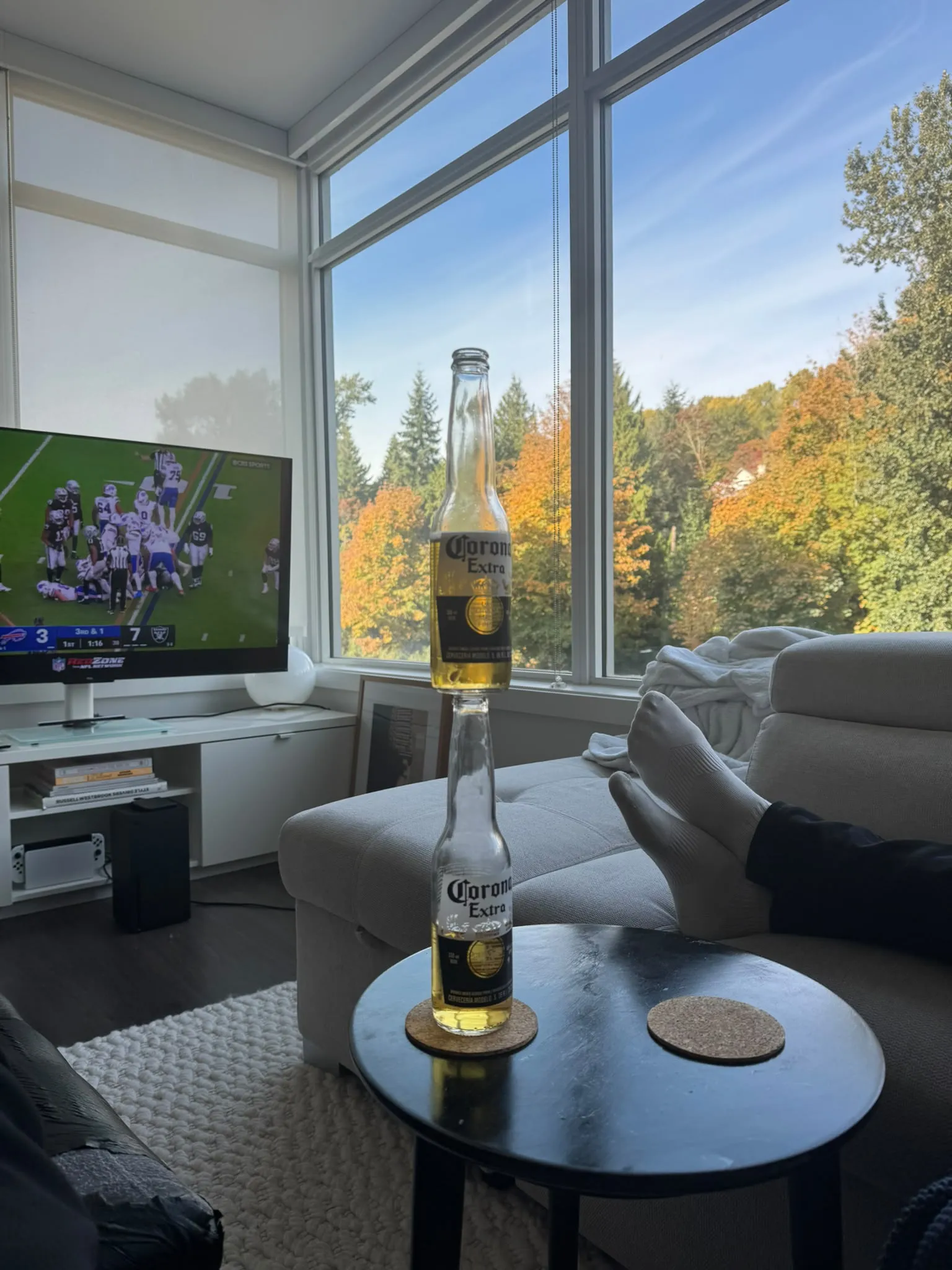 One Corona bottle balanced upright on top of another on a small round coffee table, with a football game on the TV and autumn trees outside tall windows.