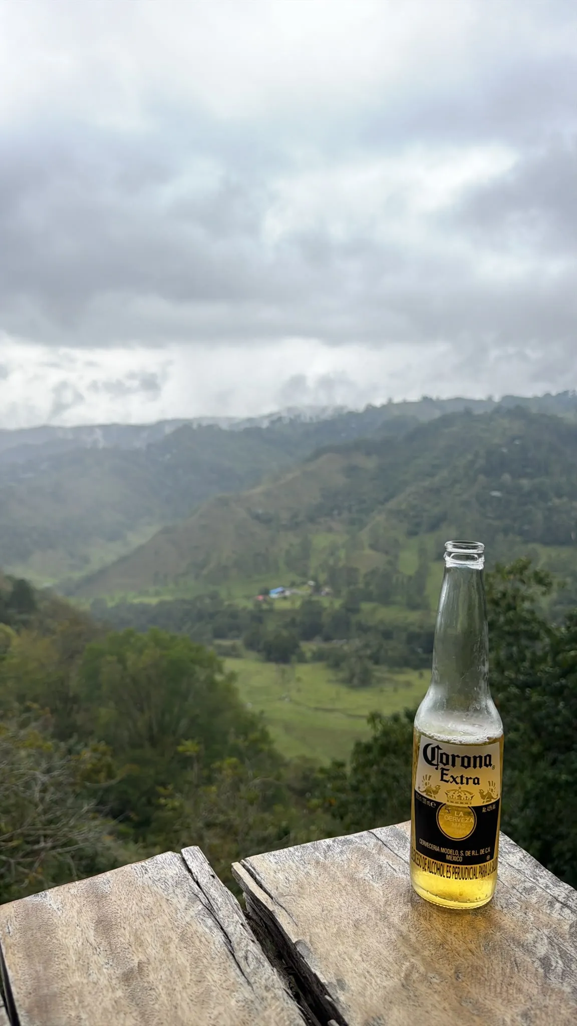 A Corona Extra bottle standing on a weathered wooden table above a green mountain valley, low cloud hanging over the hills.