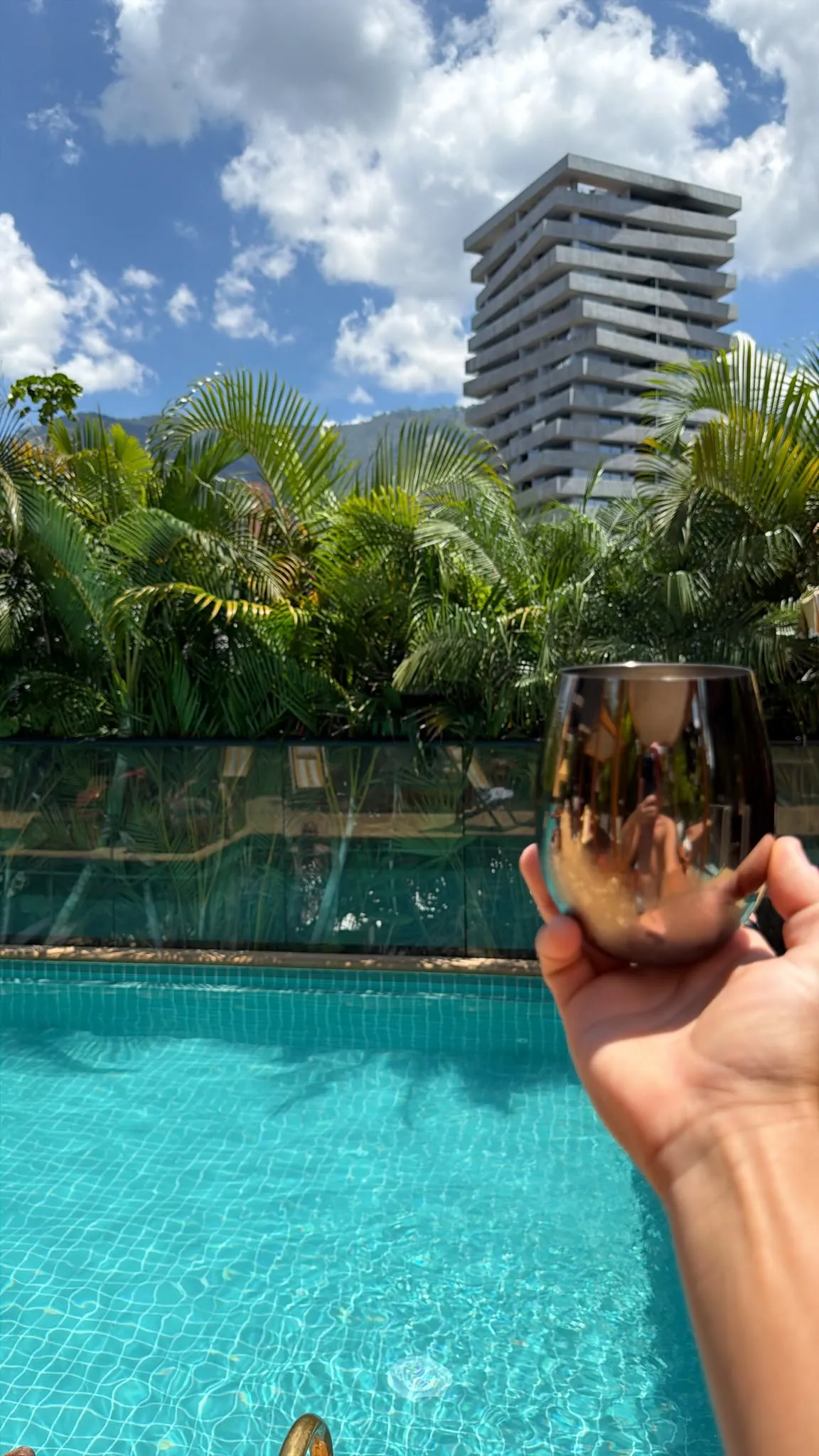 A mirrored stemless glass held up over a turquoise swimming pool, with palm trees and a tall terraced tower block against a bright cloudy sky behind.
