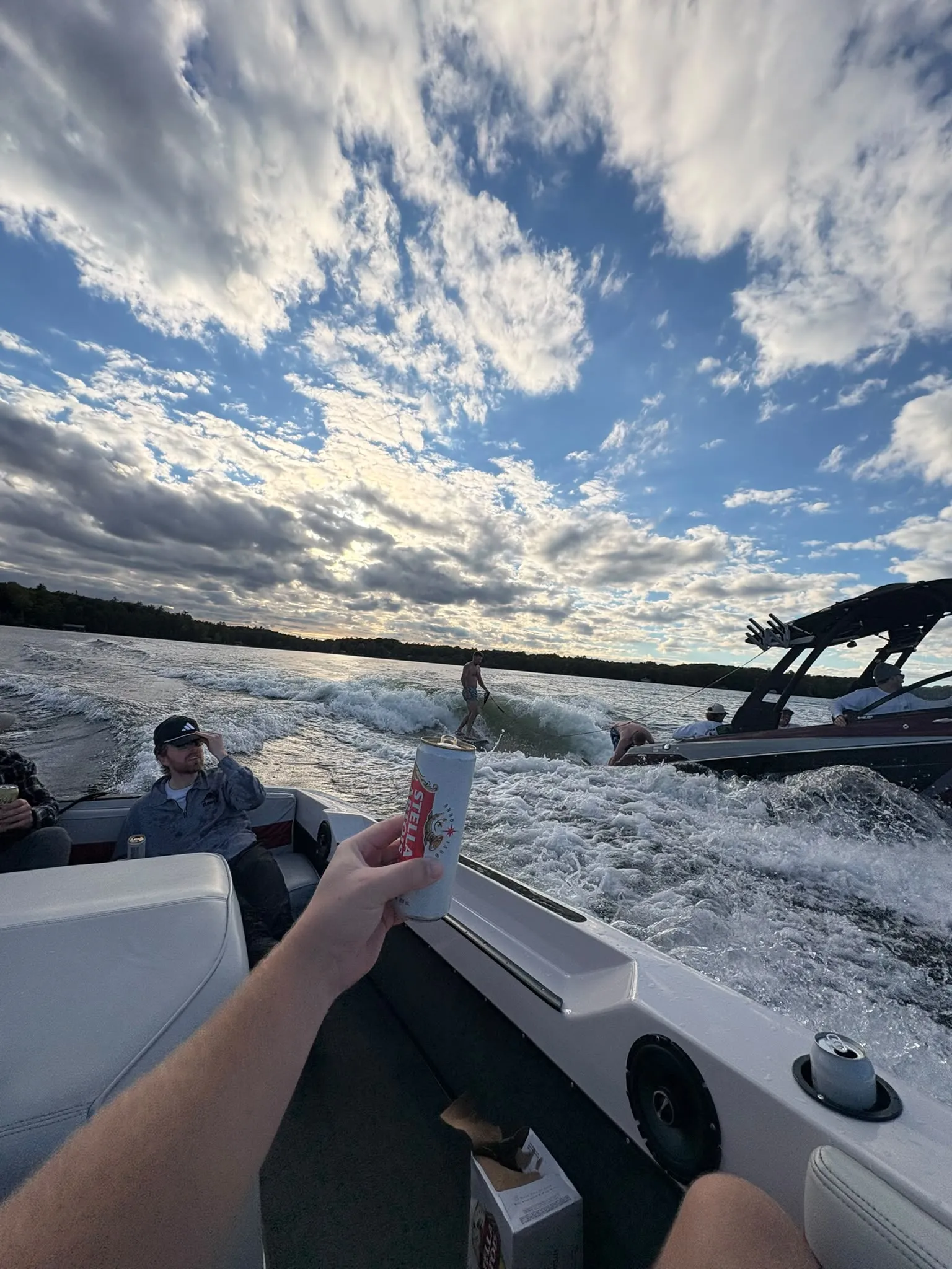 A tall can held up from the back of a moving boat as a wakesurfer rides the churning wake across a lake under dramatic sunlit clouds.